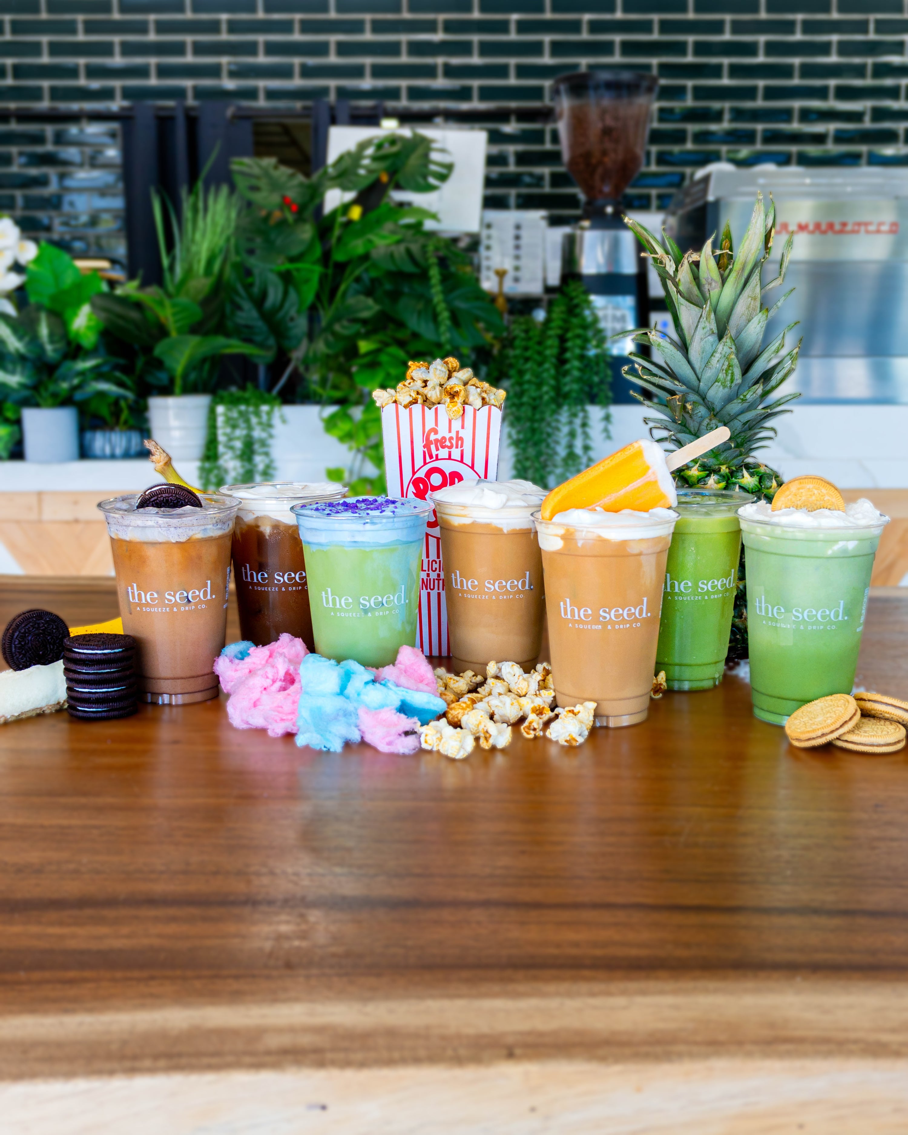 Seven colorful iced coffee and matcha drinks from 'the seed.' arranged on a wooden counter in a cafe setting, surrounded by snacks like popcorn, cookies, and a pineapple.